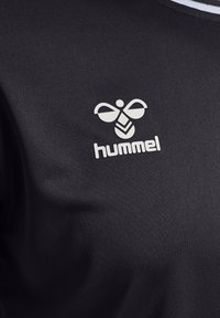 T-shirt de sport noir en tissu texturé, ornée d'un logo Hummel blanc avec un design distinctif d'abeille au centre.