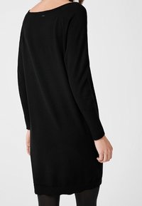Donna che indossa un lungo abito a maglia nero con maniche lunghe, in piedi con le spalle rivolte verso uno sfondo bianco semplice.