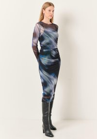 Robe à manches longues en tissu transparent bleu et noir, avec des motifs abstraits, une silhouette ajustée et un taille froncée. Associée à des bottes noires jusqu'aux genoux.