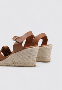 Sandalias de cuña de cuero marrón con una suela de yute estilo alpargata, con correas ajustables en el tobillo y una superficie texturizada.