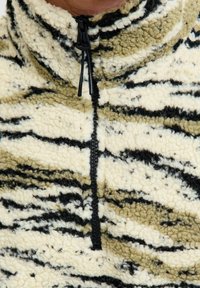 Nahaufnahme einer hellbeige und schwarz gestreiften Fleecejacke mit einem schwarzen Halbreißverschluss und Reißverschluss-Anhängern am Kragen.