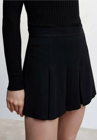 Donna che indossa una maglietta nera a coste con maniche lunghe infilata in una mini gonna plissettata nera a vita alta, in piedi con il braccio rilassato.