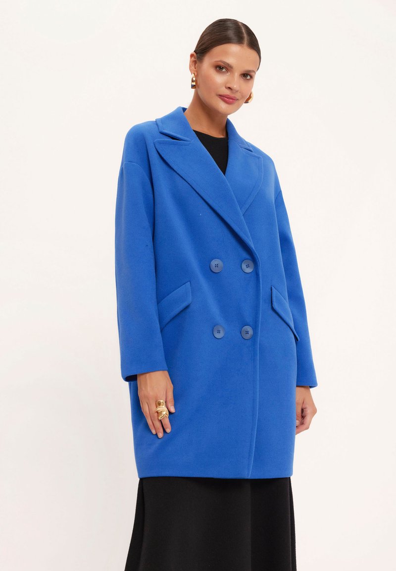 Niebieski płaszcz dwurzędowy z dużymi klapami, dwoma przednimi kieszeniami i guzikami. Gładka tekstura i oversize'owy krój.