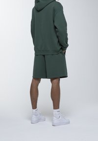 Sudadera verde con pantalones cortos a juego, ambos de mezcla de algodón. Zapatillas blancas y calcetines de longitud media. Vista trasera que muestra un ajuste relajado y un diseño minimalista.