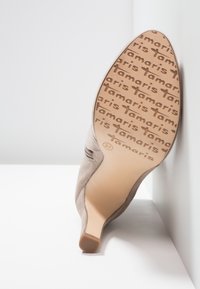 Beige heeled shoe angled to show sole with repeated "Tamaris" logo pattern and size 37 marking on a white surface.