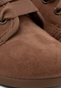 Pantofii din suede maro au o textură netedă, o formă rotunjită, curele din material textil și gaozuri metalice pe partea superioară. Talpa este din cauciuc și este căptușită.