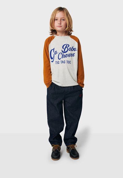 Langärmliges grau-orange Raglan-Shirt mit blauer Schrift, kombiniert mit marineblauen gestreiften Hosen. Braune Schnürschuhe runden das Outfit ab.