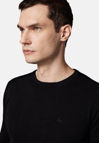 Czarny sweter z dzianiny o okrągłym dekolcie, z prążkowanymi mankietami i dołem. Subtelny detal z logo na klatce piersiowej, gładka faktura, dopasowany krój.