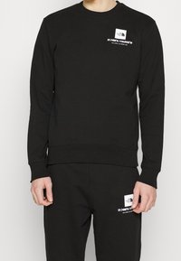 Svart sweatshirt med rund hals, ribbade ärmslut och nederkant. Noterbara grafiska detaljer inkluderar koordinater och logotyp i vitt på bröstet.