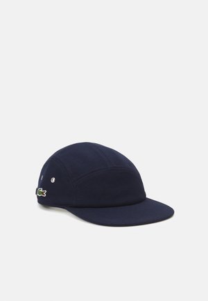 Navy blue cap made of textured fabric, featuring a flat brim, stitched panels, metal eyelets, and a small embroidered logo.