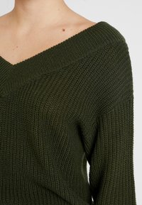 Dunkelgrüner, gestrickter Pullover mit weitem V-Ausschnitt, gerippter Textur und überschnittenen Schultern. Sichtbare Nähte als Akzent.