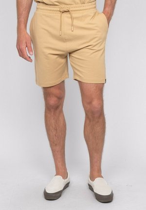 Homme portant un short beige à cordon de serrage et des chaussures blanches sans lacets, debout les mains dans les poches devant un fond clair et uni.