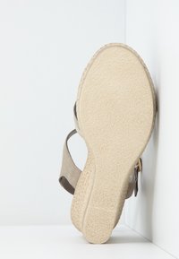 Sandale compensée avec une tige en daim beige, bride réglable et semelle en caoutchouc texturé, présentant une légère courbure au niveau du talon pour un support supplémentaire.