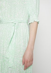 Robe vert clair avec petit motif floral blanc, ceinture à nouer à la taille, et manches courtes ballon avec bordure à volants, bras droit baissé.