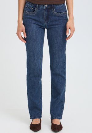 Person iført mørkeblå jeans med lige ben og dekorative syninger på lommerne, brun top og brune sko, stående mod lys baggrund.