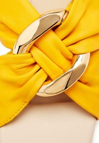 Ceinture en cuir jaune avec un design noué, dotée d'une boucle en métal doré brillant au centre, mettant en avant une texture lisse et une couleur vive.