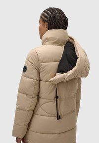 Šviesiai ruda pūkinė striukė su aukštu apykakle, siuvinėta tekstūra ir juodu vidiniu pamušalu. Papildyta virvele apatinėje dalyje ir logotipu ant rankovės.