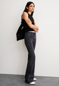 Zwarte mouwloze top, grijze flare jeans en witte hakken. Model staat met gekruiste armen en toont een gladde, gestroomlijnde textuur.