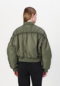 Olivgrön, vadderad bomberjacka med ribbad krage och nederkant, med en texturerad yta och överdimensionerad siluett. Svarta jeans bärs.