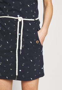 Granatowa sukienka bawełniana z białym paskiem ze sznurka, z wzorem małych ikonek związanych z plażą. Zawiera boczne kieszenie z drewnianymi akcentami.