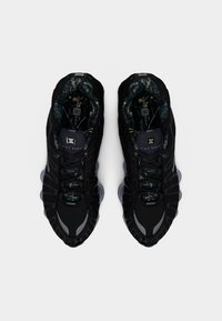 Μαύρα αθλητικά παπούτσια Nike Shox με υφασμένο πάνω μέρος, με γκρι λεπτομέρεια και εσωτερική επένδυση με σχέδιο. Η εξωτερική σόλα έχει ορατά στοιχεία απορρόφησης κραδασμών.