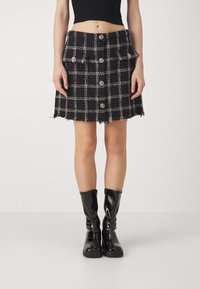 Mini-jupe à carreaux noirs avec un tissu texturé, dotée de deux poches avant, de cinq boutons et d'un ourlet effiloché. Portée avec des bottes brillantes montant jusqu'aux genoux.