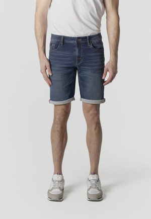 Hombre que lleva pantalones cortos de mezclilla azul arremangados, camiseta blanca y zapatillas blancas, de pie sobre un fondo claro y liso.