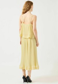 Vestido plisado de capa amarilla claro con tirantes finos y dobladillo hasta los tobillos, que presenta una textura suave y un diseño fluido. Tacones altos negros.