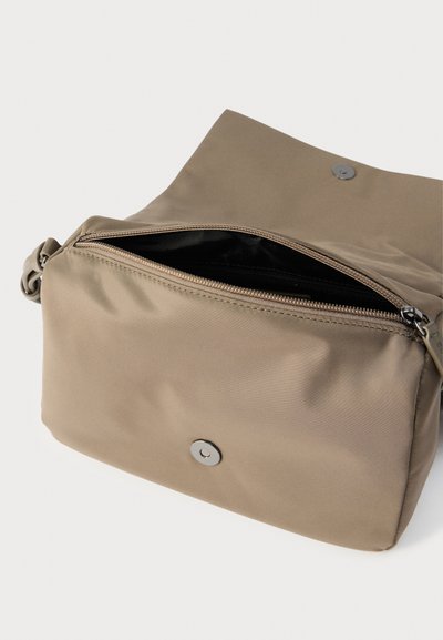 Μπεζ τσάντα ώμου με καπάκι και μεταλλικό κούμπωμα, μερικώς ανοιχτό φερμουάρ που αποκαλύπτει μαύρο εσωτερικό ύφασμα.