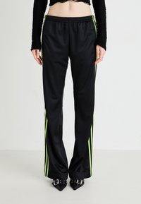 Melni satīna treniņbikses ar zaļām strīpām, elastīgu jostu un brīvu piegriezumu, kombinētas ar norādītām melnām kurpēm. Gluda tekstūra.