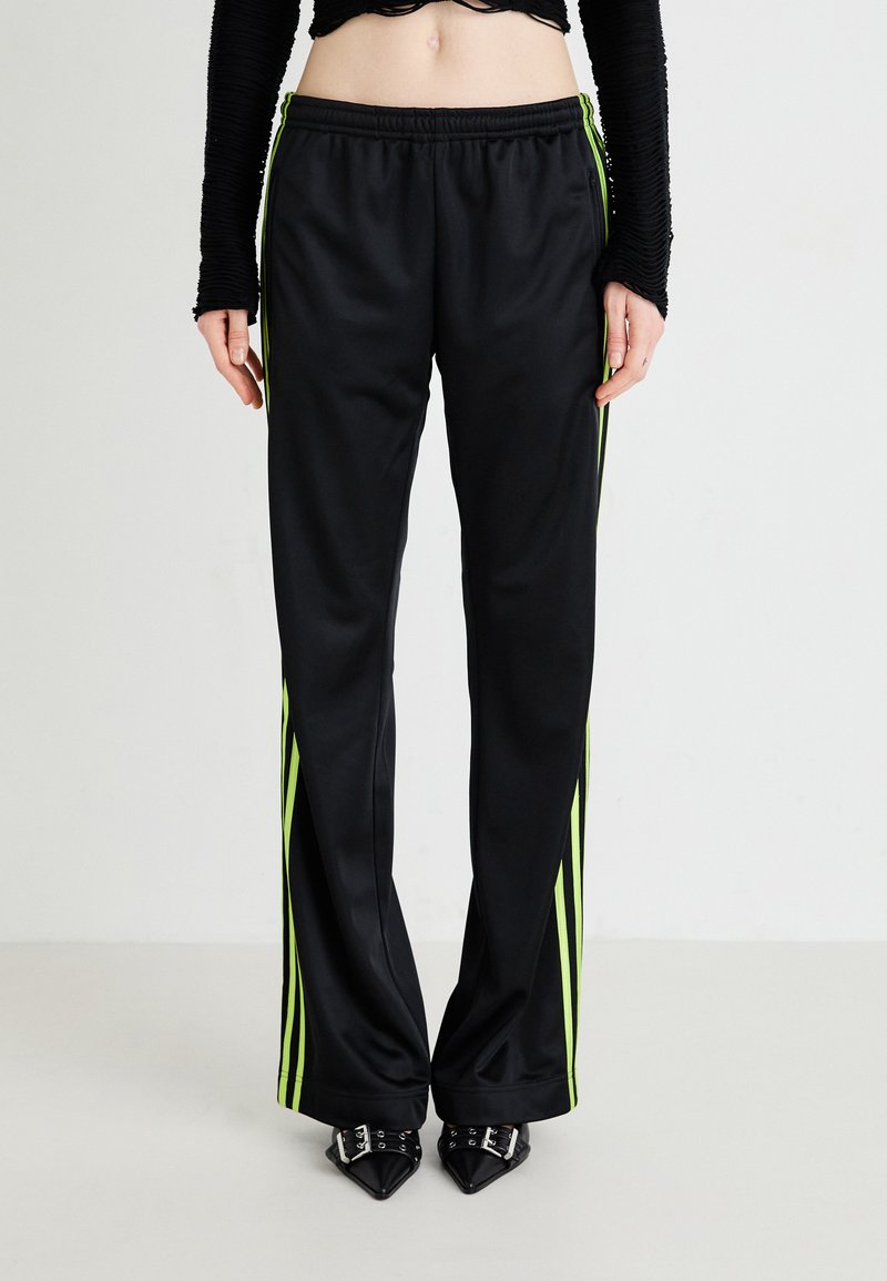 Melni satīna treniņbikses ar zaļām strīpām, elastīgu jostu un brīvu piegriezumu, kombinētas ar norādītām melnām kurpēm. Gluda tekstūra.