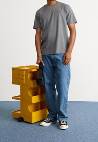 Gray cotton t-shirt, blue denim jeans, black sneakers. Standing next to a yellow, multi-shelf storage unit with circular cutouts.