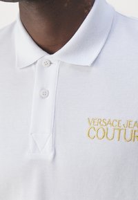 Polo blanc en tissu respirant avec un tissage texturé, comportant deux boutons et un logo "VERSACE JEANS COUTURE" brodé en or.