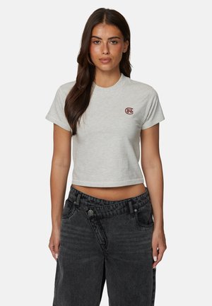 Femme aux longs cheveux foncés portant un t-shirt court gris clair avec un logo sur la poitrine et un jean noir taille haute, tournée vers l'avant.