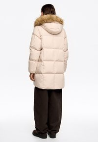 Chaqueta de plumas en un beige claro, con un ribete de piel sintética en la capucha, diseño acolchado y una longitud por debajo de las caderas. Combinada con pantalones oscuros sueltos.