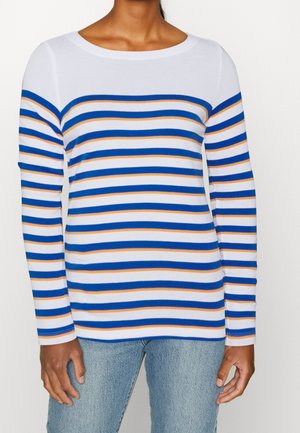 Persona con camisa blanca de manga larga con rayas horizontales en azul y naranja, combinada con jeans de mezclilla azul claro.