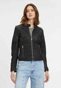 Schwarze Lederjacke mit Reißverschluss vorne, Stehkragen, vier Rei�ßverschlusstaschen und strukturierten Schulterakzenten. Tragen über einem grünen Oberteil.