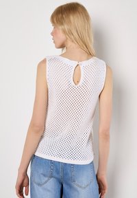 Haut sans manches en tricot blanc avec un tissage ouvert texturé et un détail en trou de serrure au dos, associé à un pantalon en denim bleu clair.