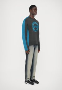 Mann steht auf einem weißen Hintergrund und trägt einen dunkelgrauen und blauen Versace Couture Jeans Pullover, ausgewaschene weit geschnittene Jeans und schwarze Sneakers.