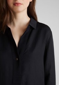 Eine Frau mit heller Haut und braunem Haar trägt ein schwarzes Hemd mit Kragen und einem sichtbaren Knopf, vor einem einfarbigen grauen Hintergrund.