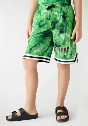 Bambino che indossa pantaloncini in rete verdi e neri con la scritta "FUTURE" e sandali neri aperti con due cinturini, in piedi su uno sfondo bianco.