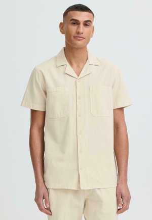 Homme portant une chemise beige à manches courtes boutonnée avec deux poches poitrine et un pantalon beige assorti, debout devant un fond clair uni.