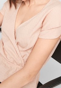 Blusa rosa claro con un escote en V profundo, mangas cortas y tejido texturizado que presenta detalles fruncidos en la cintura.