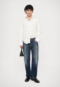 Λευκό πουκάμισο με κουμπιά, σκούρο μπλε τζιν με ίσια γραμμή, μαύρες δερμάτινες παπούτσια, κρατώντας μια μικρή μαύρη τσάντα. Απλό φόντο.