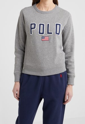 Femme portant un sweat-shirt gris avec "POLO" et un patch du drapeau américain, associé à un pantalon bleu marine avec un petit logo rouge.