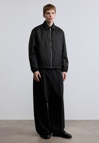 Μαύρο μπουφάν τύπου μπόμβερ με υφή, ασημένιο φερμουάρ και ελαστικές μανσέτες, συνδυασμένο με μαύρο παντελόνι φαρδιάς γραμμής και μαύρα παπούτσια.