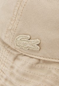 Tan cotton cap with textured fabric, featuring a stitched beige logo of a crocodile on the side. Curved brim and visible seam detailing.