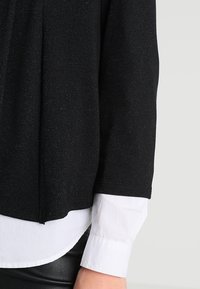 Svart stickad topp med en texturerad yta, med en kontrasterande vit skjorta under och en rak fåll. Långa ärmar.