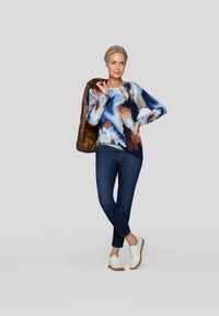 Langärmliges Oberteil mit blauen, braunen und weißen abstrakten Mustern, kombiniert mit dunklen Jeans und weißen Sneakers. Strukturiierte braune Jacke über dem Arm getragen.