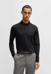 Schwarzes Langarm-Polohemd mit einem mittelhohen Kragen und drei Knöpfen, kombiniert mit hellgrauen Hosen und einem schwarzen Gürtel mit einer silbernen Schnalle.